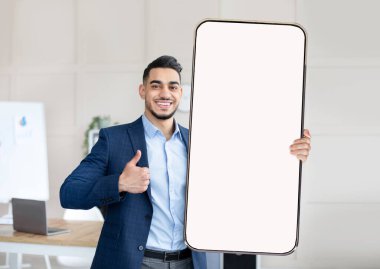 Arap başarılı işadamı boş akıllı telefon gösterisi yapıyor ve başparmağını kaldırıyor, uygulama ya da online teklif öneriyor