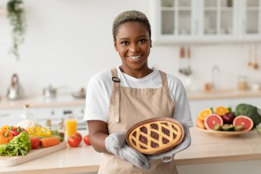 Gülümseyen binlerce yıllık Afro-Amerikan önlüklü kadın minimalist mutfağın içinde lezzetli turtalar gösteriyor.