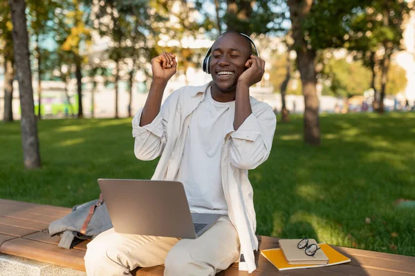 Радостный африканский студент-мужчина, отдыхающий в кампусе с ноутбуком, в наушниках и наслаждающийся музыкой на открытом воздухе — стоковое фото
