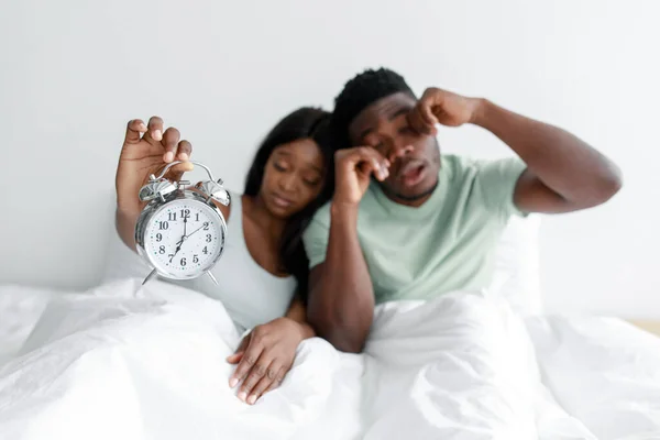 Uykulu Afro-Amerikan kadın ve erkek uyanıyor, gözlerini ovalıyor, yatak odasındaki beyaz yatağa oturuyor ve çalar saati gösteriyor. — Stok fotoğraf