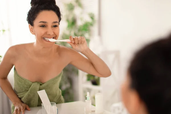 Wanita muda yang cantik dengan senyum yang indah mengenakan handuk setelah mandi, menyikat gigi dengan sikat gigi di dekat cermin di rumah — Stok Foto