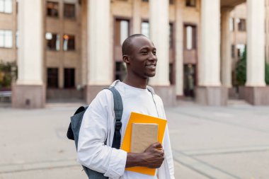 Sırt çantası ve çalışma kitapları olan gülümseyen Afrikalı Amerikalı erkek öğrenci üniversite binasının arka planında poz veriyor.