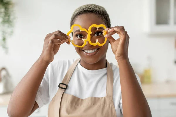 Felice millenaria donna afroamericana casalinga in grembiule divertirsi e mette pezzi di pepe agli occhi come occhiali — Foto Stock