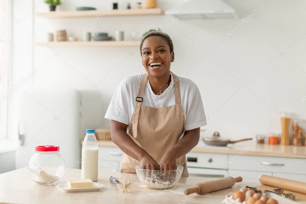 Laughing young african american lady in apron making pie dough in minimalist kitchen interior