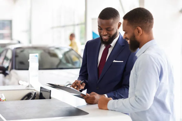 Homem preto alegre que compra o carro, assinando papéis com vendedor no escritório da concessionária — Fotografia de Stock