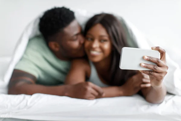 Χαμογελώντας χιλιετή αφροαμερικανός άνδρας φιλιά γυναίκα, κυρία λήψη selfie στο smartphone σε λευκό κρεβάτι — Φωτογραφία Αρχείου