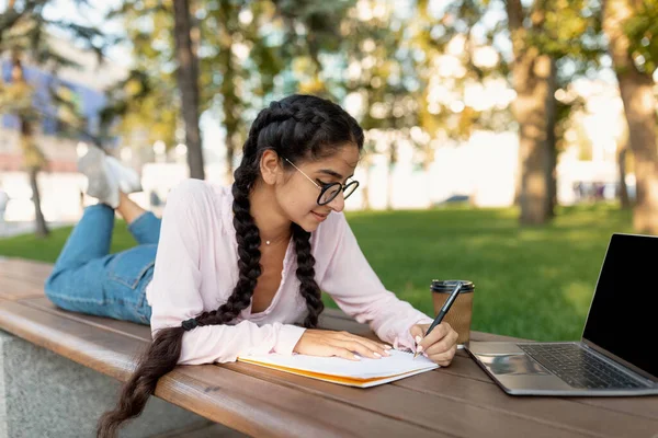 Indien étudiant fille prendre des notes tout en apprenant à l'extérieur, couché sur le banc avec ordinateur portable, passer du temps dans le parc du campus universitaire — Photo