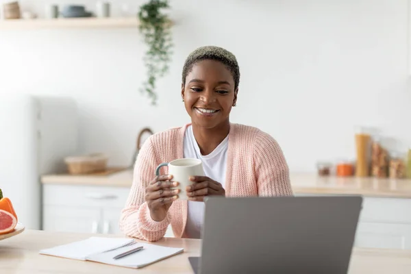Happy millennial Afrikaans amerikaanse vrouw hold cup en werken met laptop in moderne minimalistische keuken interieur — Stockfoto