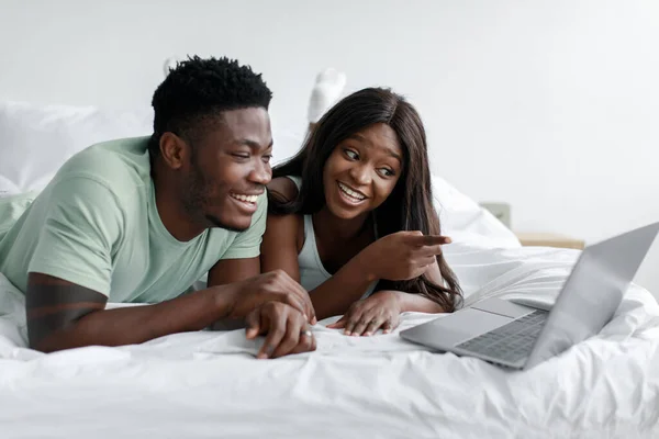Feliz riendo joven marido negro y la esposa miran a la computadora portátil, tienen videollamada en la cama en el dormitorio blanco —  Fotos de Stock