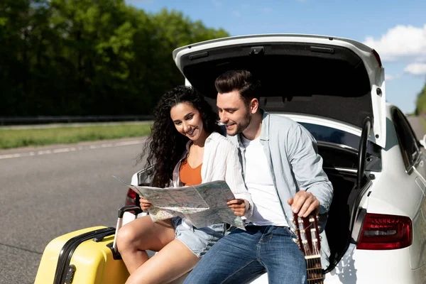 Positives Paar auf Autoreise, Landkarte ansehen, Pause machen, gemeinsam im Kofferraum sitzen — Stockfoto