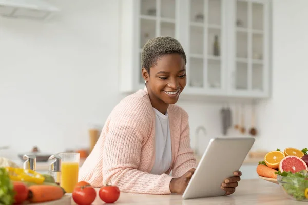 Smiling millennial african american lady blogger looking at tablet at table with organic fruits — Stock Photo, Image