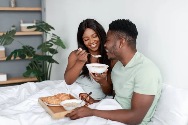 Souriant millénaire afro-américaine femme se réveiller, nourrir mari flocons d'avoine au lit dans la chambre à coucher lumière intérieur — Photo