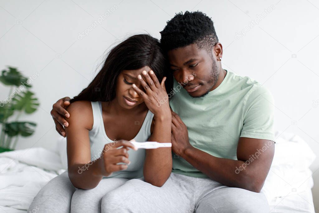 Depressed sad young black female and male upset of negative pregnancy test result in light bedroom interior