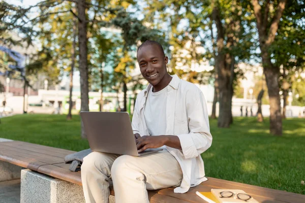 Descanse no campus. Feliz afro-americano estudante universitário masculino relaxante ao ar livre com laptop, sentado no banco no parque — Fotografia de Stock