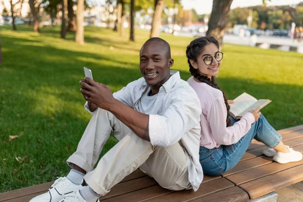 Happy diverse college students sitting back to back, using smartphone and book, resting outdoors on bench in park — Stock Photo, Image