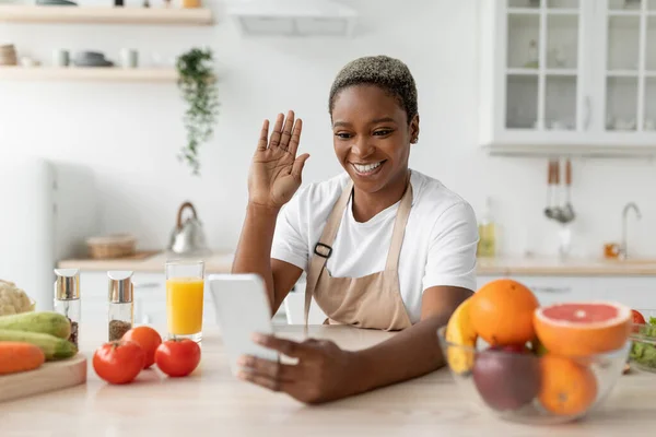 천 년의 흑인 여성 이 앞치마를 두르고 전화기로 손을 흔들며 식탁에 앉아 야채와 과일 과 함께 부엌에서 — 스톡 사진