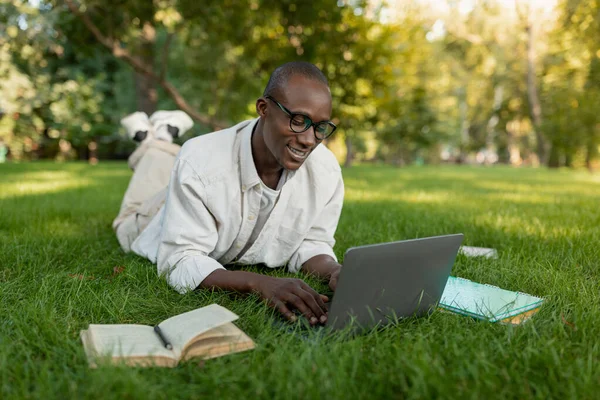 Веселый чернокожий студент, пользующийся ноутбуком, изучая интернет на открытом воздухе, лежит на траве в студенческом городке — стоковое фото