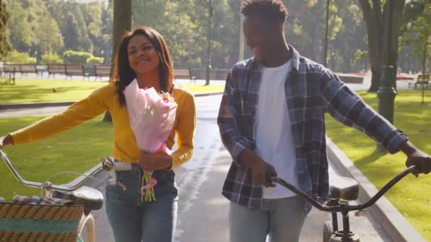 Kedves fiatal afro-amerikai férfi és nő séta kerékpárok keresztül nyilvános park, kövesse lövés, lassított felvétel — Stock videók