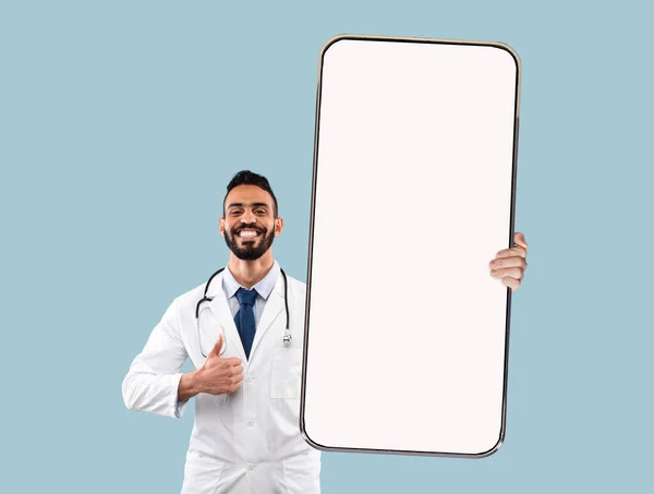 Árabe médico masculino mostrando smartphone com tela branca em branco e gesto polegar sobre fundo azul, mockup — Fotografia de Stock