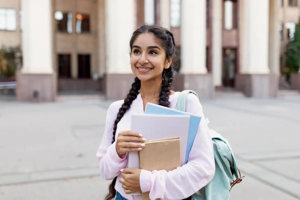 Retrato ao ar livre de estudante indiana alegre com mochila e cadernos de trabalho em pé perto do prédio da faculdade — Fotografia de Stock