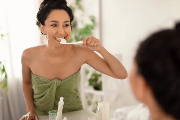 Mujer morena bastante joven con hermosa sonrisa cepillarse los dientes delante del espejo en casa, espacio libre — Foto de Stock