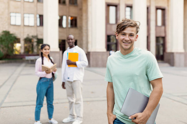 Happy multiracial university students posing outdoors near university building, selective focus on caucasian guy