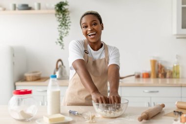 Önlüklü mutlu siyah kadın hamur yapıyor, çağdaş hafif mutfakta turta pişiriyor.