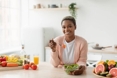 Minimalist mutfak iç mekanında organik sebzelerle salata yiyen neşeli Afro-Amerikan kadını.