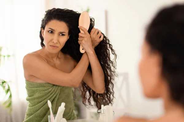 Mujer joven trastornada que tiene problemas para cepillarse el cabello enredado cerca del espejo en casa, espacio libre —  Fotos de Stock