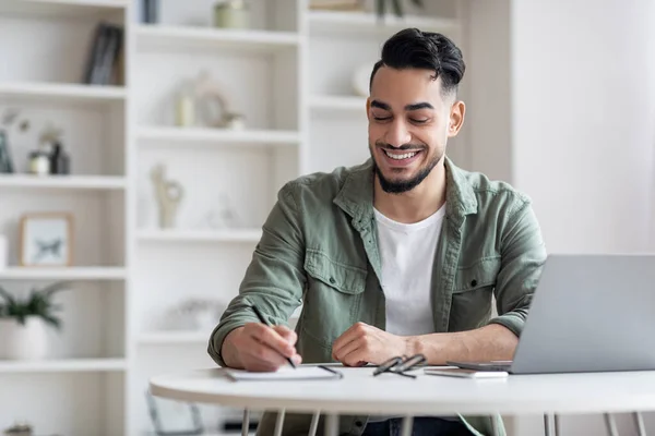 Lächelnder arabischer Mann macht sich Notizen, während er im Home Office mit Laptop arbeitet — Stockfoto