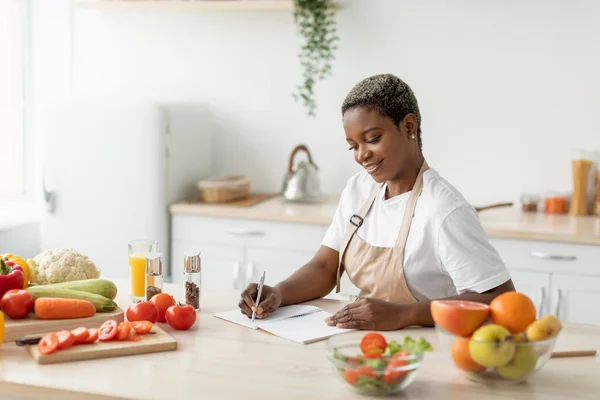 Cheerful millennial black lady in apron makes notes on table with organic vegetables and fruits — Stock Photo, Image