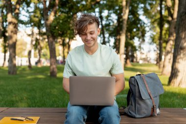 Bilgisayarlı neşeli öğrenci ev ödevi yapıyor ya da online derse katılıyor, dışarıda bankta oturuyor.