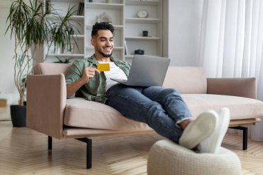 Çevrimiçi ödemeler. Gülümseyen Arap evde dizüstü bilgisayar ve kredi kartı kullanıyor.