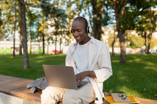 Χαμογελώντας μαύρο άντρα χαλαρώνοντας με φορητό υπολογιστή και ακουστικά στο πάρκο μετά τα μαθήματα, απολαμβάνοντας το χρόνο που περνούν σε εξωτερικούς χώρους — Φωτογραφία Αρχείου
