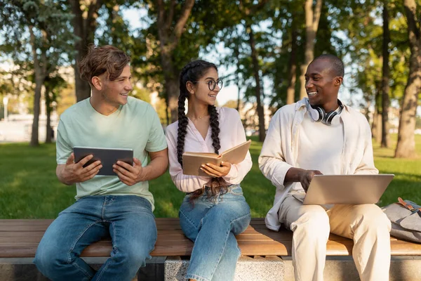 Три однокласники коледжу готуються до лекцій разом, сидячи на лавці в університетському містечку з книгами та пристроями — стокове фото