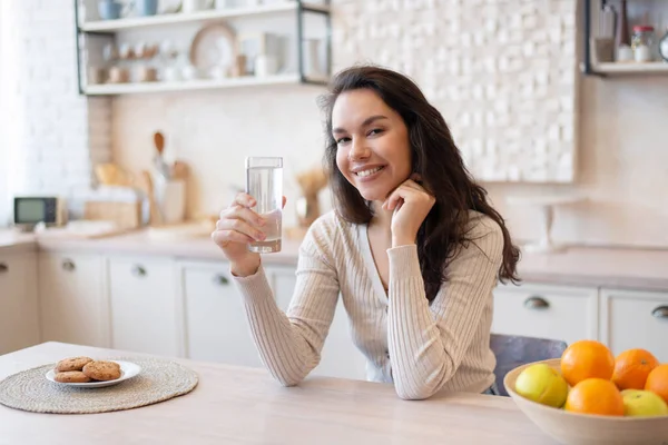 Porträt einer positiven Frau, die Wasser aus Glas trinkt, am Tisch in der Küche sitzt und in die Kamera lächelt — Stockfoto