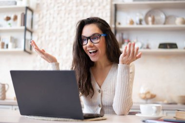 Heyecanlı kadın portresi çevrimiçi video görüşmesi yapıyor, iş arkadaşlarıyla ya da müşterileriyle dizüstü bilgisayarla konuşuyor, mutfakta oturuyor.