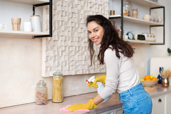 Szczęśliwa gospodyni domowa w gumowych rękawiczkach sprzątanie tabeli w kuchni i uśmiecha się do kamery, utrzymując jej dom schludny i czysty — Zdjęcie stockowe