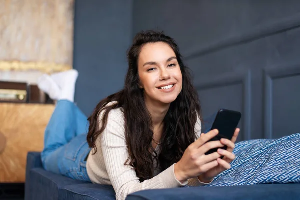 Portret van een vrolijke dame die op bed ligt en lacht naar de camera terwijl ze thuis kletst met een geliefde of surft op sociale media — Stockfoto