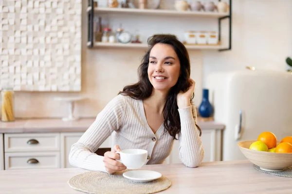 Mladá dáma s šálkem kávy pití oblíbený nápoj v lehkém kuchyňském interiéru, dívá se jinam a usmívá — Stock fotografie