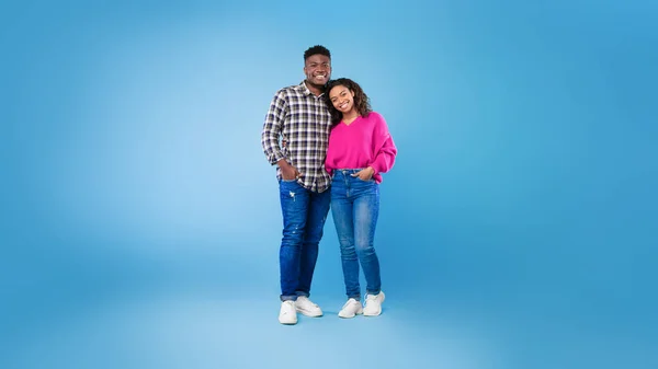 Полная длина счастливой молодой афроамериканской пары в повседневной одежде, обнимающей и смотрящей в камеру на синем фоне — стоковое фото