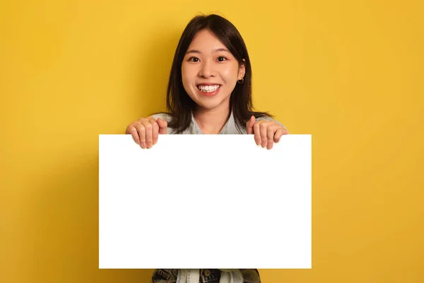 Feliz joven asiática sosteniendo pancarta de papel en blanco, sonriendo a la cámara en el fondo amarillo del estudio, maqueta —  Fotos de Stock