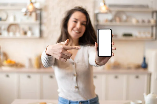 Donna positiva mostrando schermo smartphone vuoto e puntando su di esso mentre in piedi in cucina, messa a fuoco selettiva, mockup — Foto Stock