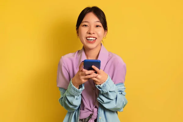 Mulher asiática milenar alegre usando telefone celular, conversando na web, trabalhando ou estudando on-line no fundo amarelo — Fotografia de Stock