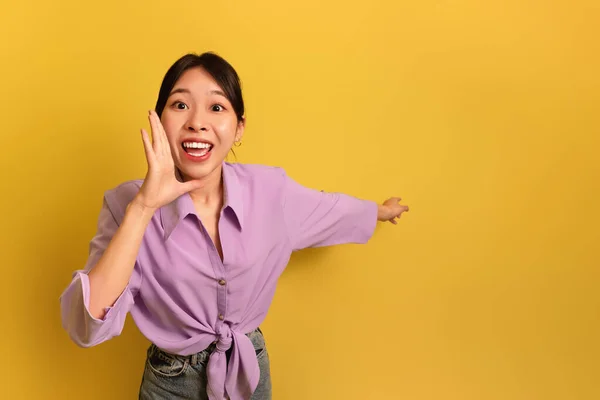 Jovem mulher asiática excitada gritando OMG e apontando para o espaço em branco no fundo do estúdio amarelo — Fotografia de Stock