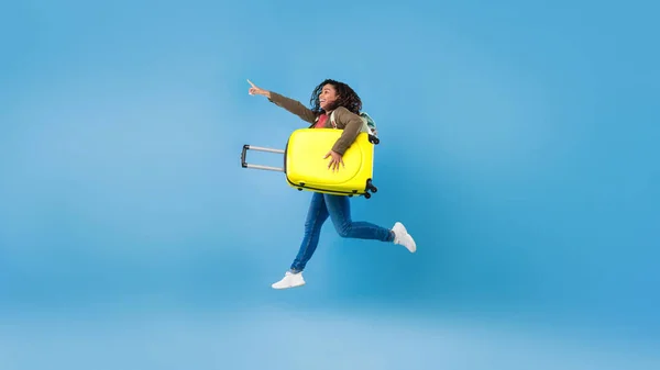 Щаслива молода чорна жінка стрибає з валізою і вказує на порожнє місце для реклами туристичних агентств на синьому фоні — стокове фото