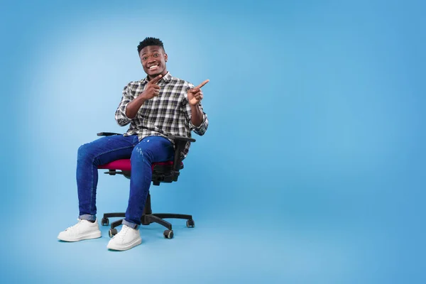 Emocionado jovem negro sentado na cadeira, apontando dois dedos de lado, oferecendo espaço para o seu anúncio — Fotografia de Stock
