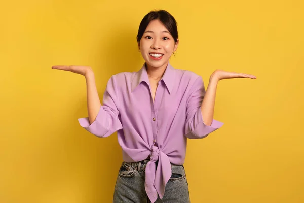 Mulher asiática feliz comparando opções, fazendo escalas com as mãos, segurando objetos invisíveis no fundo amarelo, mockup — Fotografia de Stock