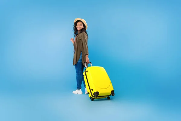Full length portrait of happy black female traveler walking with suitcase over blue studio background, copy space — Stock Photo, Image
