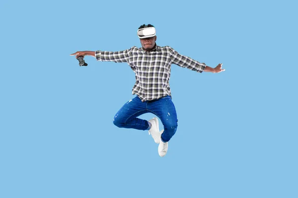 Joven afroamericano con auriculares VR sosteniendo joystick, saltando en el aire sobre fondo azul —  Fotos de Stock